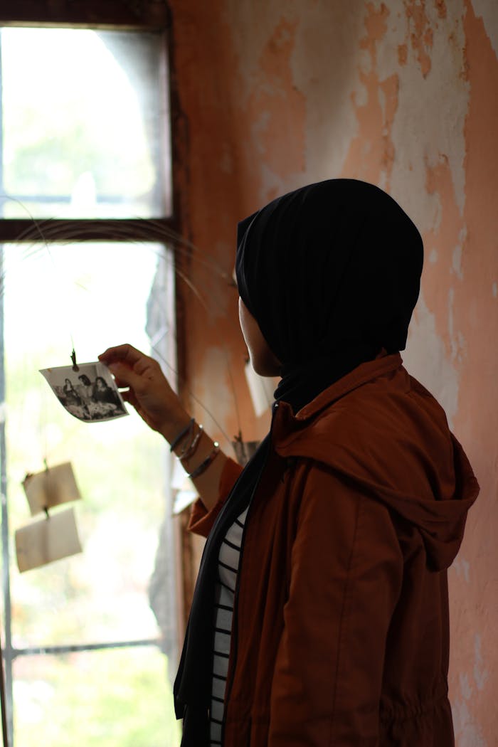 A woman in a hijab holding a photo, reflecting on memories indoors. Captured in soft lighting.